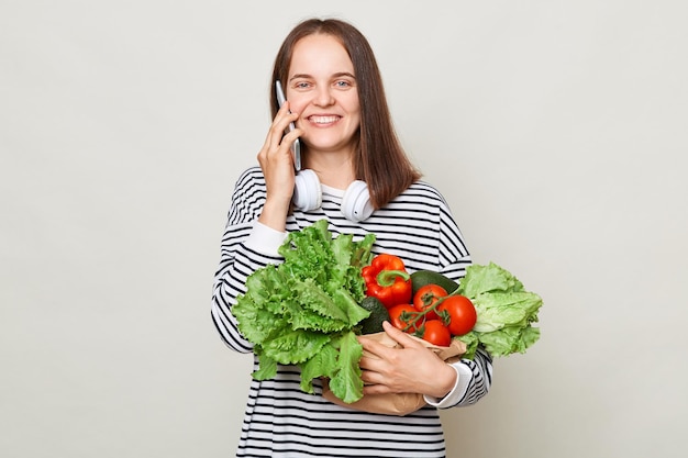 Légumes frais du marché Mode de vie végétarien sain Enthousiaste joyeuse femme aux cheveux bruns parlant téléphone portable achetant des légumes frais posant sur fond gris regardant la caméra