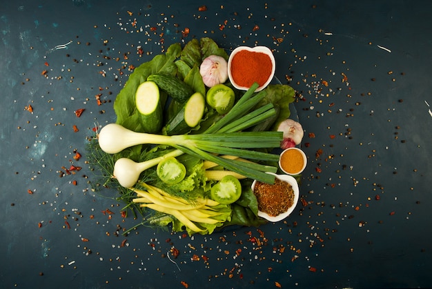 Légumes frais et épices sur une planche de bois ronde sur une pierre sombre. LE CONCEPT DE MILLESIME. LES ÉPICES DU CONCOMBRE DE TOMATES VERTES JUS D'ASPERGES DE MAÏS SUR UNE SURFACE FONCÉE.