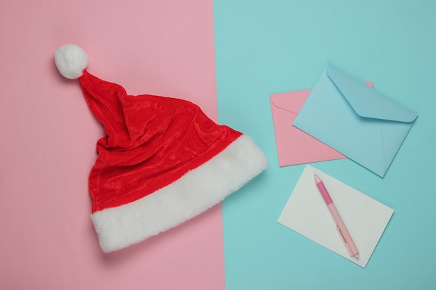 Lettre au Père Noël. Bonnet de Noel, enveloppe avec une lettre et un stylo sur fond bleu rose. Noël à plat. Vue de dessus