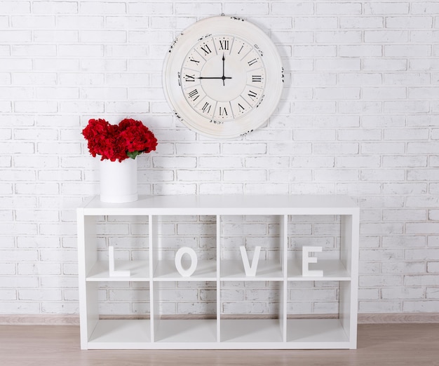 Lettres en bois formant le mot AMOUR dans une étagère moderne, des fleurs et une horloge vintage sur un mur de briques blanches