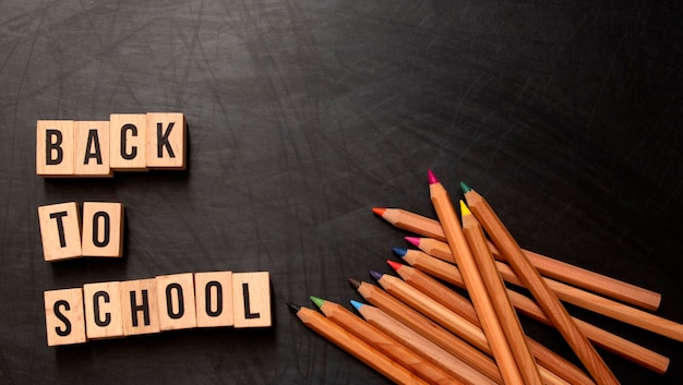 Lettres carrées en bois avec la phrase "Retour à l'école" écrite en anglais sur un fond de tableau noir et à côté de crayons de couleur