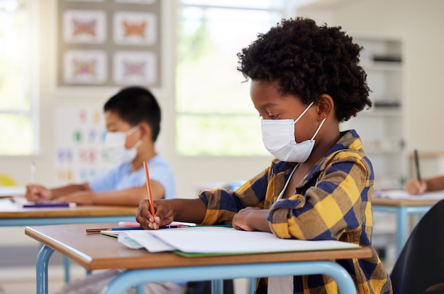 Élève en classe pendant la pandémie de covid pour apprendre l'éducation et étudier avec un masque pour la protection de la sécurité et le protocole Petit enfant de la maternelle ou du primaire écrivant dans un livre au bureau