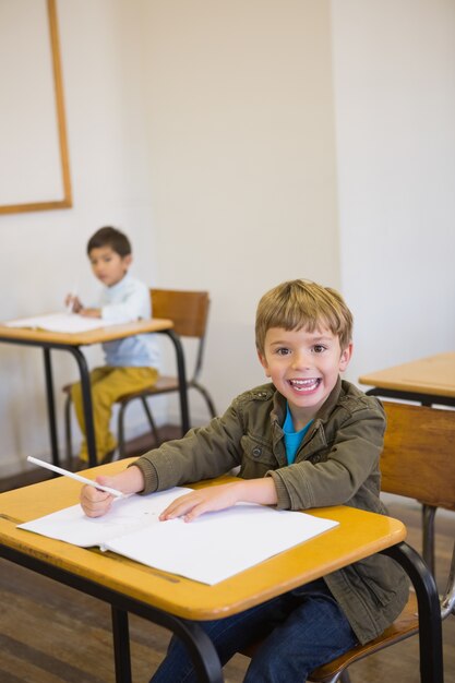 Élève écrit dans le bloc-notes à son bureau, souriant à la caméra