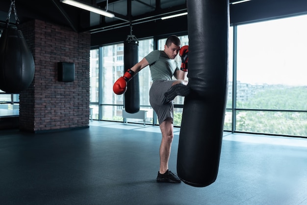 Lève la jambe. Boxer professionnel expérimenté aux cheveux noirs battant le sac de boxe avec la jambe tout en s'entraînant dur