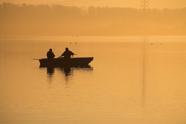 Lever de soleil orange sur le lac Silhouettes de deux pêcheurs dans un bateau brouillard à l'aube