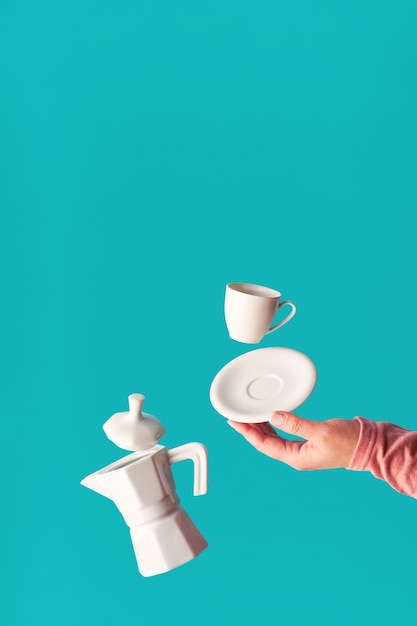 Lévitation à la mode Grains de café volants, tasse à expresso avec soucoupe en équilibre sur l'index de la main féminine. Lévitation de cafetière en céramique. Mur de café bleu menthe tendance, espace pour votre texte.