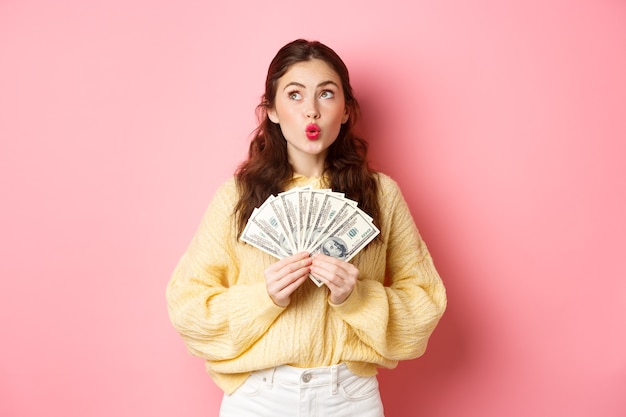 Les lèvres plissées d'une jeune femme réfléchie, regardant de côté avec un visage pensif, tenant des billets d'un dollar, rêvant d'acheter quelque chose avec de l'argent, debout sur fond rose