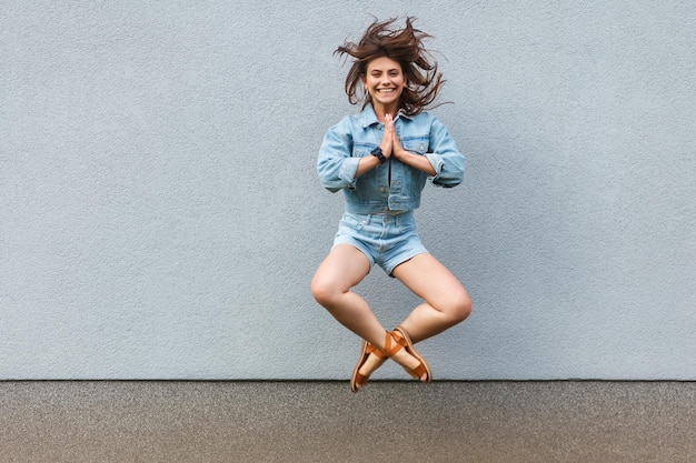 Libre heureuse belle femme en jeans décontractés style denim en été sautant au mur bleu clair avec des mains de palmier namaste et regardant la caméra avec un sourire à pleines dents. concept de bonheur, de liberté et de réussite.