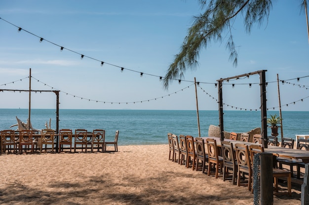 Lieu de célébration avec table longue en bois, chaise, ampoule suspendue et décoration végétale de style rétro sur la plage en mer tropicale