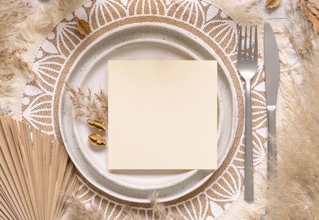 Lieu de table de mariage avec carte vierge carrée sur plaque sur la vue de dessus de napperon bohème. Maquette de carte de mariage Boho. Modèle beige tropical avec plantes séchées, feuilles de palmier et herbe de la pampa, placez le texte pour le texte