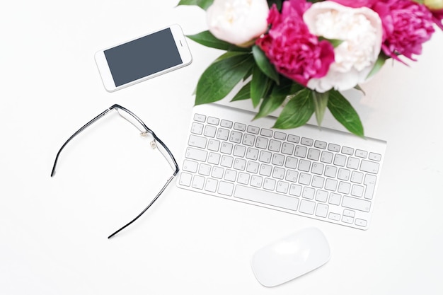 Lieu de travail avec clavier, lunettes, ordinateur portable, téléphone portable et fleurs de pivoine roses et blanches sur fond de tableau blanc. Mise à plat, vue de dessus