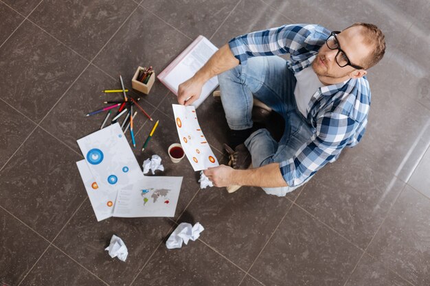 Lieu de travail inhabituel. Sérieux bel homme intelligent assis sur le sol et tenant son dessin tout en pensant à son nouveau projet