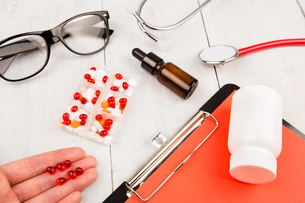 Lieu de travail d'un médecin Pilules à la main presse-papiers stéthoscope rouge et verres sur table en bois blanc