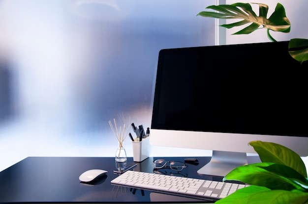 Photo lieu de travail avec ordinateur moderne sur une table en verre, écran noir simulé, plante d'intérieur et fournitures.
