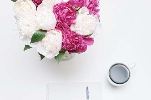 Lieu de travail avec tasse de café, papier, stylo et fleurs de pivoine rose et blanche sur fond de tableau blanc. Mise à plat et vue de dessus