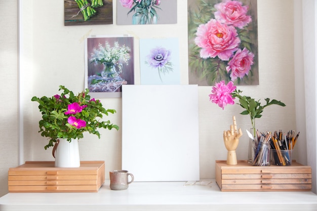 Lieu de travail vide de femme artiste à côté de matériaux pour dessiner des pinceaux, des crayons et de belles peintures de fleurs roses sur le mur. Place pour le texte. Toile blanche propre