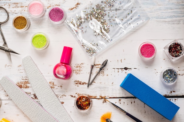 Photo limes à ongles, ciseaux, pinces, paillettes et vernis à ongles sur un fond en bois blanc.