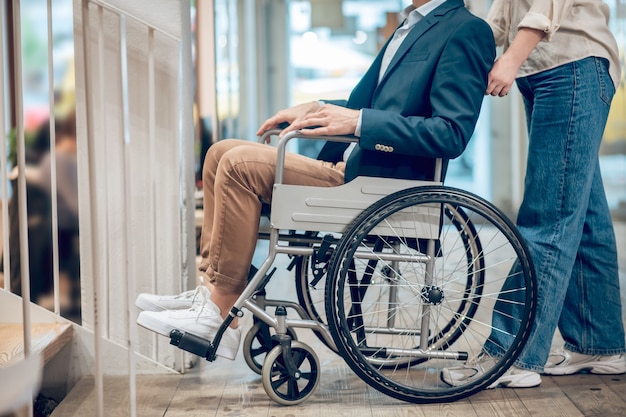 Limitation de mouvement. Homme handicapé en fauteuil roulant et assistant accompagnateur debout à proximité dans la chambre, le visage n'est pas visible