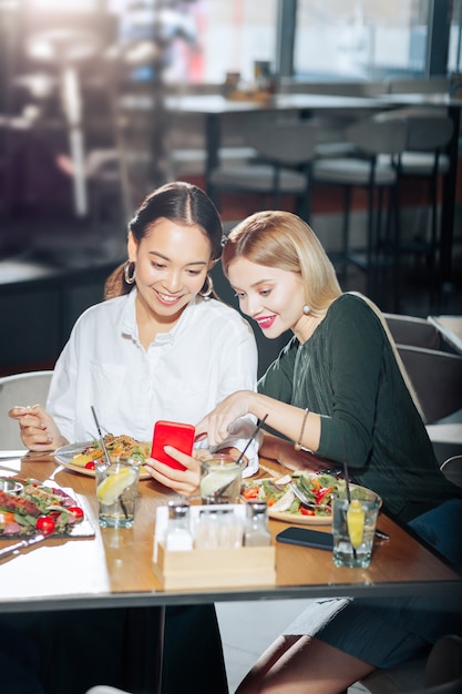 Lire le fil d'actualité Deux meilleurs amis lisant un fil d'actualité curieux sur un smartphone rouge pendant le dîner