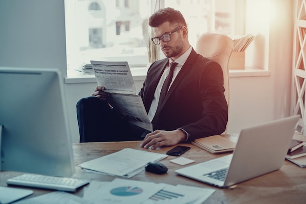 Lire un journal frais. Jeune homme sérieux en tenue de soirée lisant le journal alors qu'il était assis au bureau