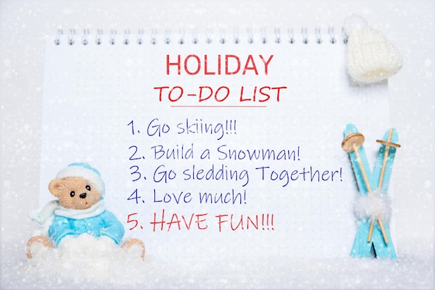 Liste de choses à faire pour les vacances. Bloc-notes avec liste de choses à faire : skier, faire un bonhomme de neige, faire de la luge, aimer, s'amuser et un ours en peluche en vêtements bleus, des skis bleus, un chapeau blanc sur de la neige blanche et des flocons de neige