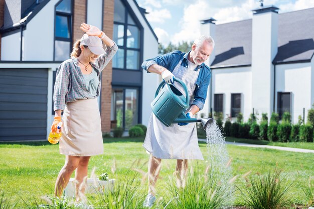 Lit d'arrosage. Mari et femme portant des vêtements décontractés et des tabliers se sentant charmants et heureux tout en arrosant leur lit de jardin