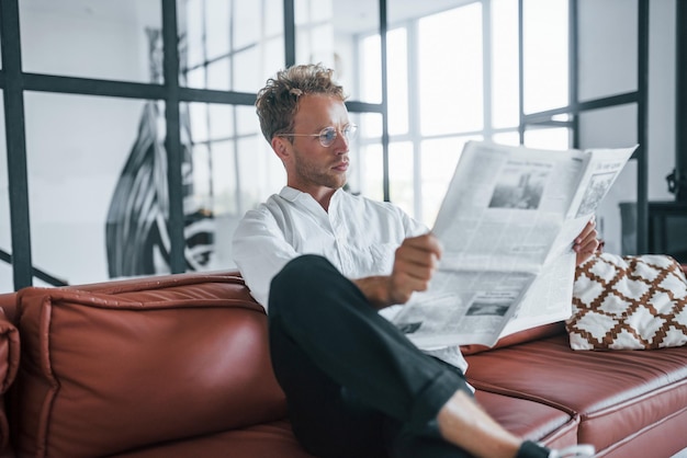 Lit le journal Caucasien jeune homme en chemise blanche élégante à l'intérieur à la maison