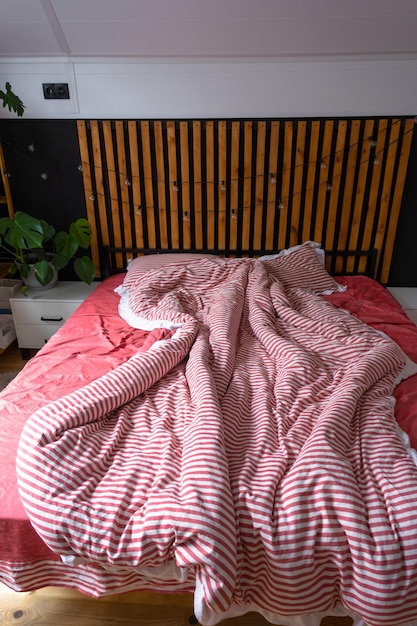 Lit non fait avec du linge rayé rouge et un désordre dans la chambre à coucher de style Loft mur noir intérieur avec des lames de bois lit métallique plantes en pot sur une fenêtre trapèze dans le grenier moderne maison verte