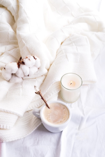 Literie avec un plaid moelleux et une tasse de café, des fleurs en coton et une bougie.