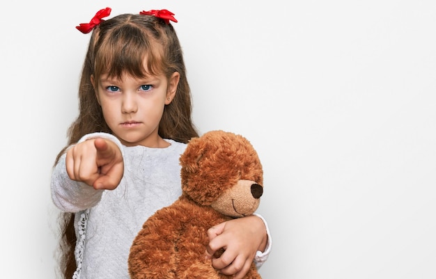 Little Caucasian girl kid hugging teddy bear animal en peluche pointant du doigt vers l'appareil photo et à vous geste confiant à la grave