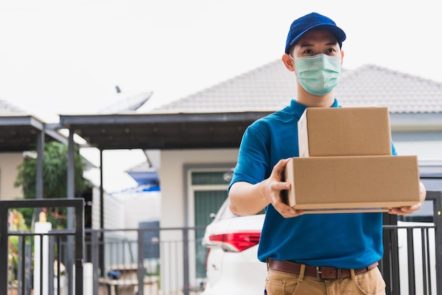 Livraison asiatique courrier express jeune homme donnant des boîtes à une cliente qu'il porte un masque de protection à l'avant de la maison, sous couvre-feu coronavirus pandémique de quarantaine COVID-19