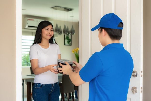 Livraison asiatique jeune homme en uniforme bleu sourire et tenant des tas de boîtes en carton devant la maison