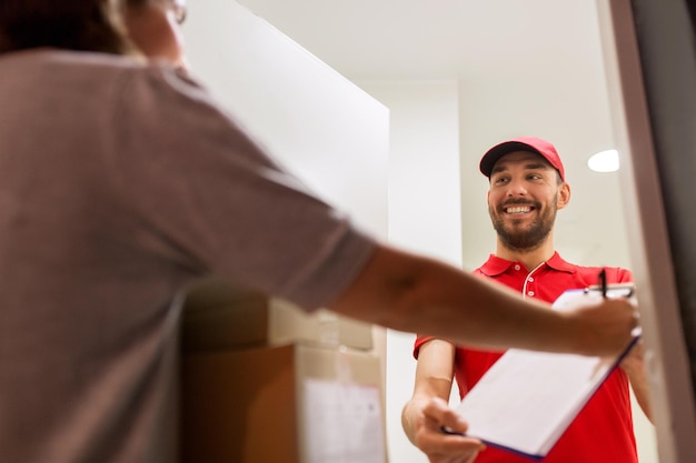 livraison, courrier, personnes et concept d'expédition - homme heureux avec clipboard livrant des boîtes de colis au client signant le formulaire à domicile