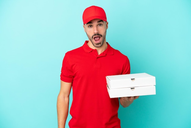 Livraison homme caucasien ramassant des boîtes à pizza isolées sur fond bleu avec une expression faciale surprise