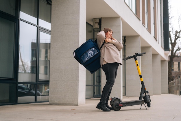 Livraison de nourriture par coursier féminin mettant un sac à dos thermo sur les épaules et montant sur un scooter électrique dans la rue de la ville