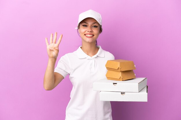 Livraison de pizza Fille russe avec uniforme de travail ramassant des boîtes de pizza et des hamburgers isolés sur fond violet heureux et comptant quatre avec les doigts