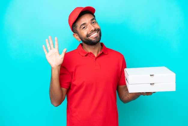 Livraison de pizza Homme brésilien avec uniforme de travail ramassant des boîtes de pizza isolées sur fond bleu saluant avec la main avec une expression heureuse