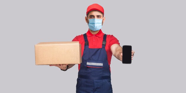 Livraison en quarantaine. Ceci est pour vous! Jeune homme avec masque médical chirurgical en uniforme bleu et t-shirt rouge debout, tenant une boîte en carton et montrant un écran de téléphone intelligent sur fond gris.