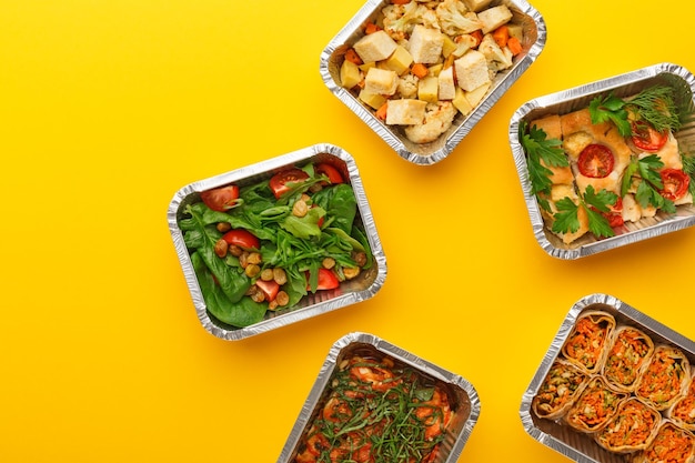 Livraison de repas sains. Casserole, rouleaux végétariens, salades de légumes frais et cuits au four dans des boîtes à emporter en aluminium sur fond jaune. Manger le bon concept, copier l'espace, vue de dessus.