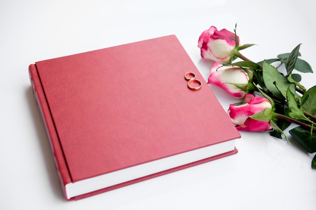 Livre ou album de mariage recouvert de cuir rouge avec deux anneaux de mariage et trois roses se trouve sur blanc