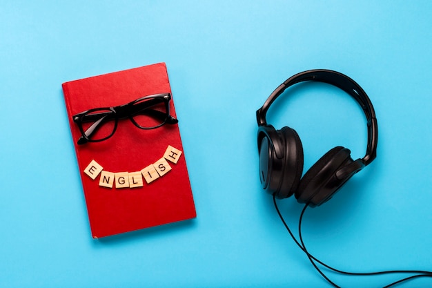 Livre avec une couverture rouge avec texte anglais, lunettes et casque noir sur fond bleu. Concept de livres audio, d'auto-éducation et d'apprentissage de l'anglais indépendamment. Mise à plat, vue de dessus