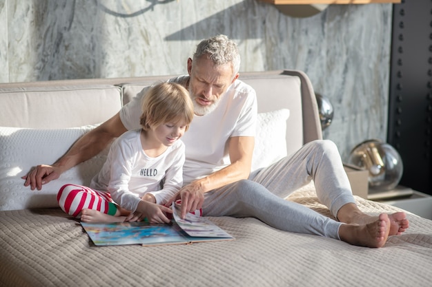 Livre divertissant. Papa barbu aux cheveux gris avec enfant souriant assis pieds nus sur le lit en regardant livre