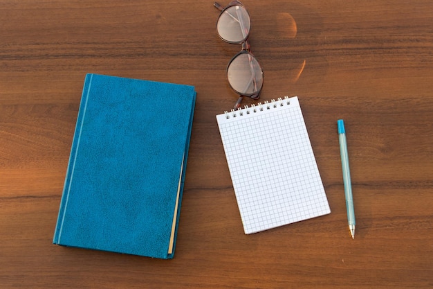 Livre fermé, lunettes, bloc-notes et stylo sur fond de bois