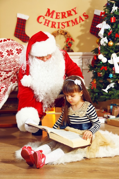 Livre de lecture du Père Noël avec une petite fille mignonne près de la cheminée et de l'arbre de Noël à la maison