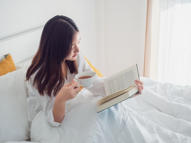 Livre de lecture de femme asiatique et de boire du café sur le lit le matin