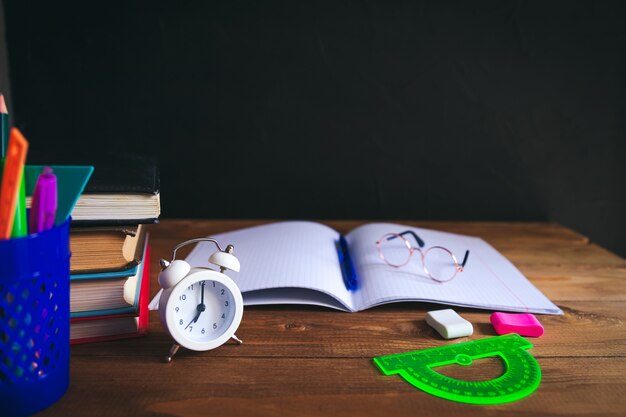 Livres, matériel d'écriture, lunettes, cahier et réveil sur le bureau. Fond noir. Concept d'apprentissage et d'auto-éducation. Mise au point sélective.