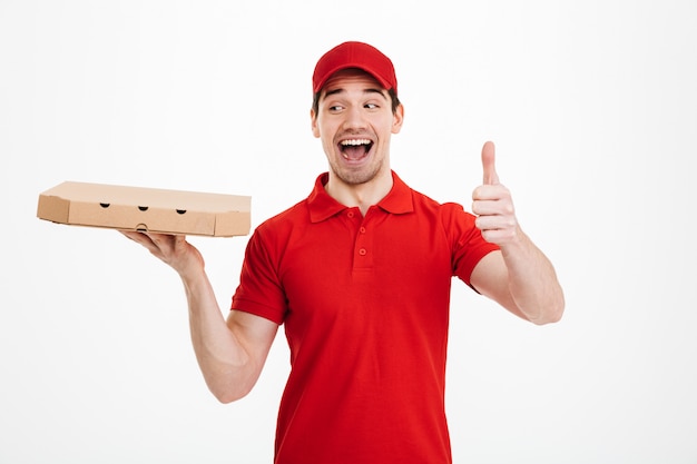 Livreur 25 ans en t-shirt rouge et casquette tenant la boîte de carton à emporter avec pizza et le pouce vers le haut avec un sourire parfait, isolé sur un espace blanc