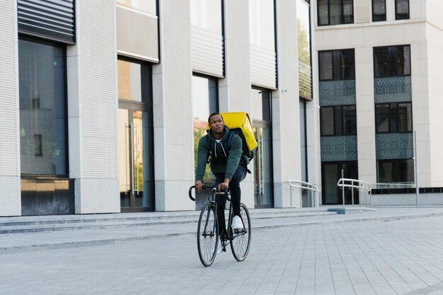 Un livreur afro-américain avec un sac à dos jaune fait du vélo dans le contexte d'un centre d'affaires moderne