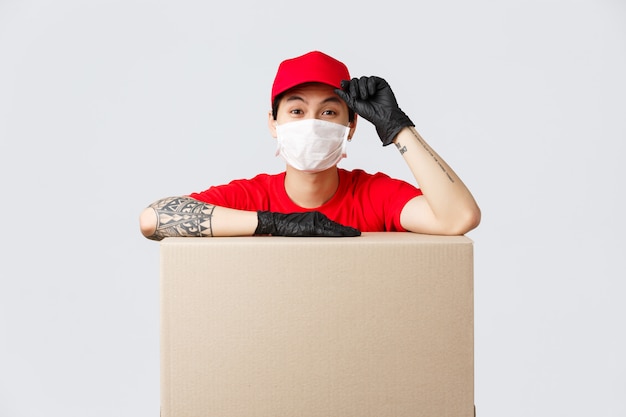 Livreur asiatique en bonnet rouge et t-shirt, service de messagerie s'appuyant sur une boîte en carton ou un colis client, saluant comme faisant la commande, transfère les marchandises aux personnes qui restent en sécurité quarantaine