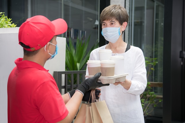 Livreur asiatique portant un masque et des gants en uniforme rouge offrant un sac de nourriture et de boisson au destinataire